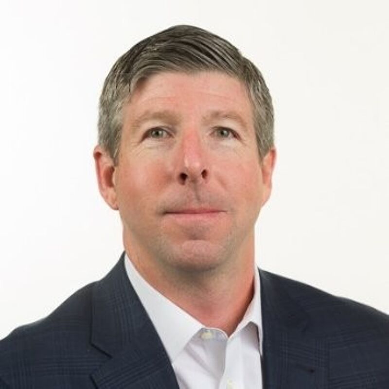 The image shows a middle-aged man with short, graying hair and a neutral expression. He is wearing a dark blue suit jacket over a white dress shirt. The background is plain white, which makes the man the focal point of the image. The lighting is bright and even, suggesting a professional headshot.